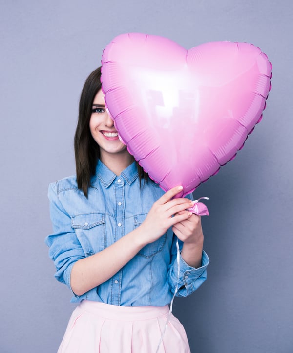 Smiling woman covering her eye with heart shaped balloon over gray background. Looking at camera