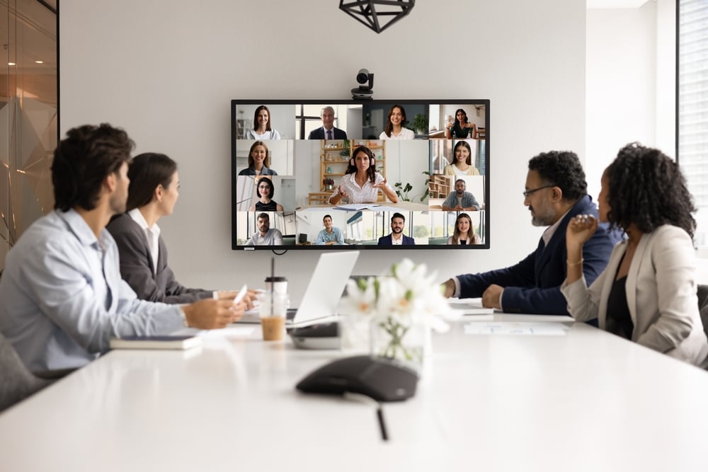 A hybrid business meeting with professionals in a conference room video conferencing remote participants on a large screen.